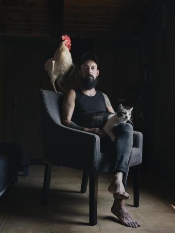 Retrato de Familia de Mon, sentado en una butaca, con el gato Levi encima y el gallo Levi sobre el respaldo.